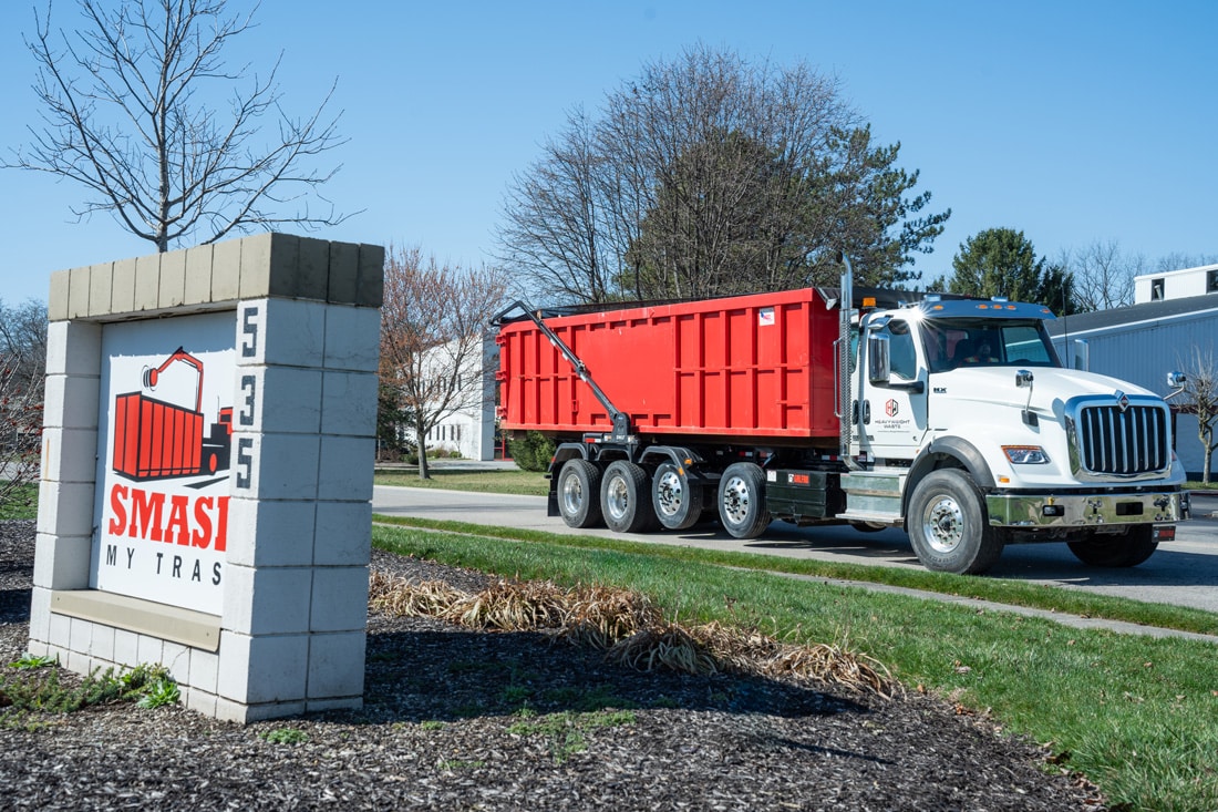 Red roll-off dumpster truck for waste removal parked beside SMASH company sign at commercial property on clear sunny day