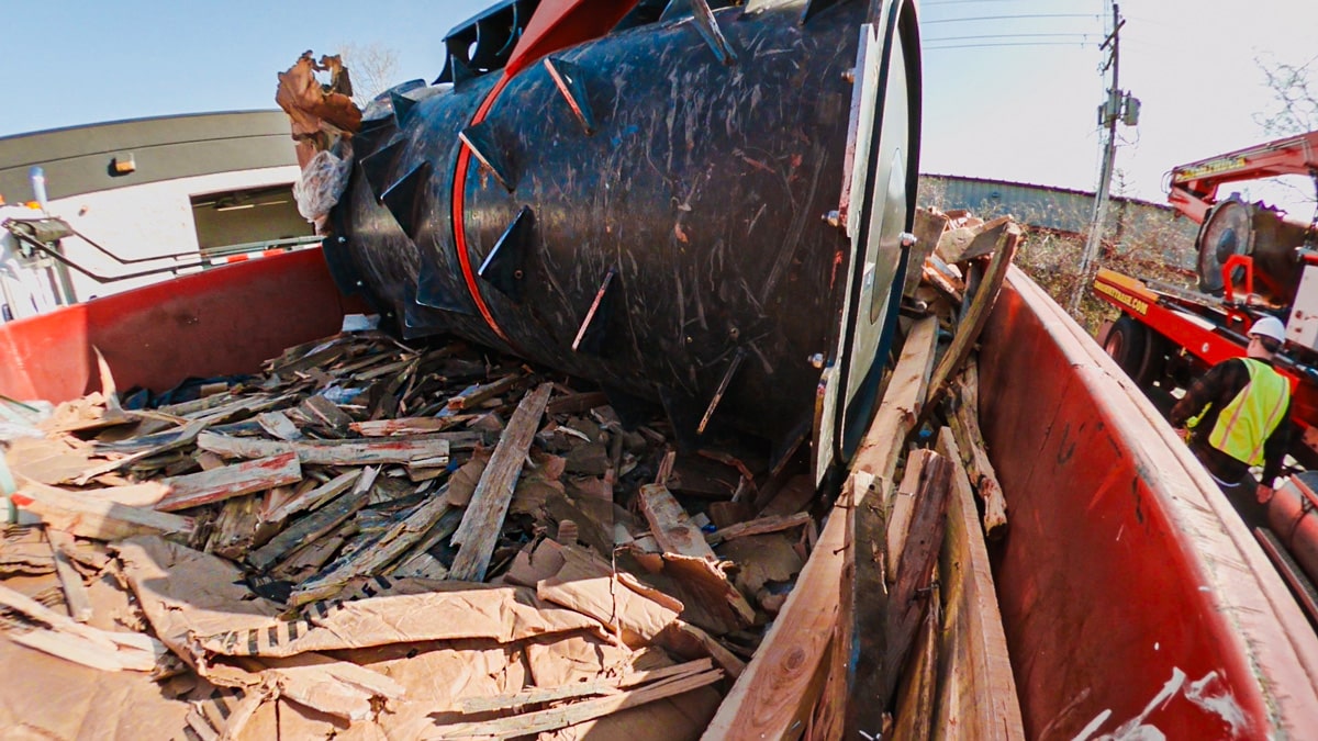 Large black industrial drum and construction debris in red dumpster with workers in safety vests nearby