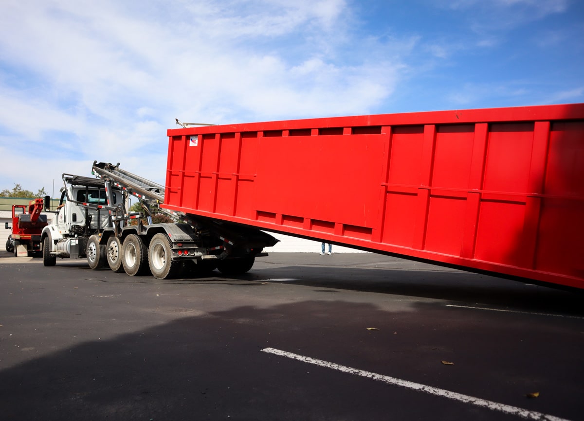 Red roll-off dumpster being loaded onto white hauling truck in a parking lot under a blue sky