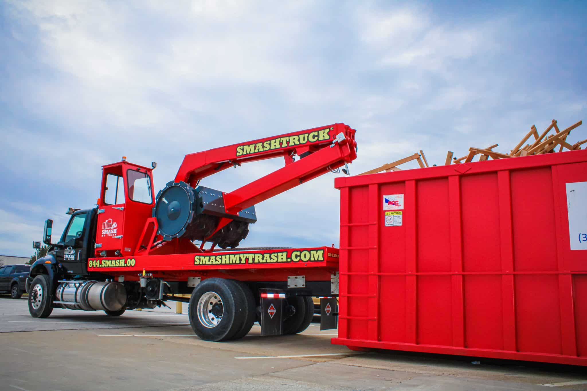 Red SmashTruck compacting construction debris into large red roll-off dumpster at a job site under cloudy sky
